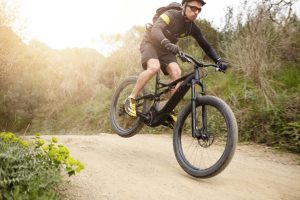 Cyclist in action jumping high on black electric motor-powered bicycle down trail in woods. Young rider wearing glasses and helmet making extreme biking stunt on e-bike while exercising outdoors